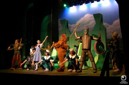 Imagem da representação do clássico O Mágico de Oz no teatro, com alguns dos personagens em cena, como o Homem de Lata, Dorothy, Espantalho, entre outros.